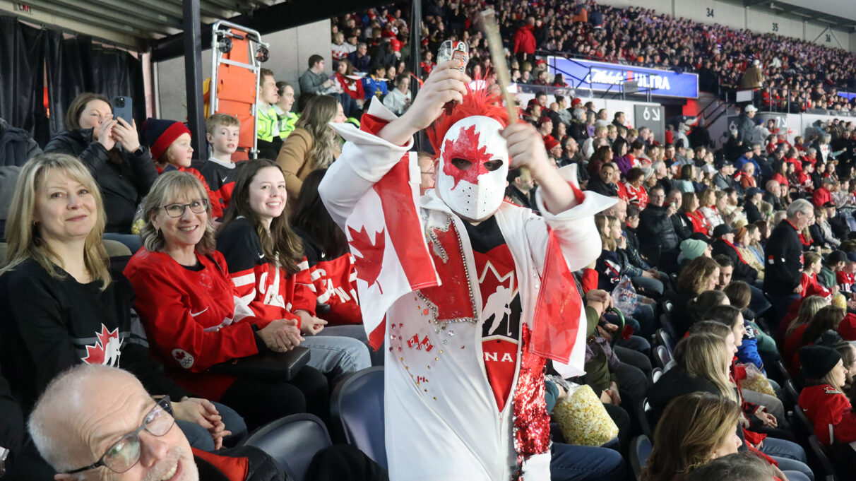 A Team Canada fan in an elaborate costume drums on a cowbell in the stands.