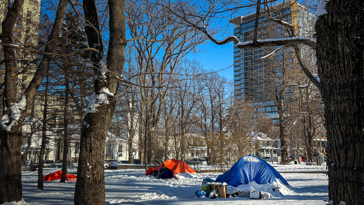 A blue tent and orange tent sit beneath the trees and on the snow in a park beside houses with a new apartment building almost finished in the background. The sky is clear and blue and there are no leaves on the brown trees.