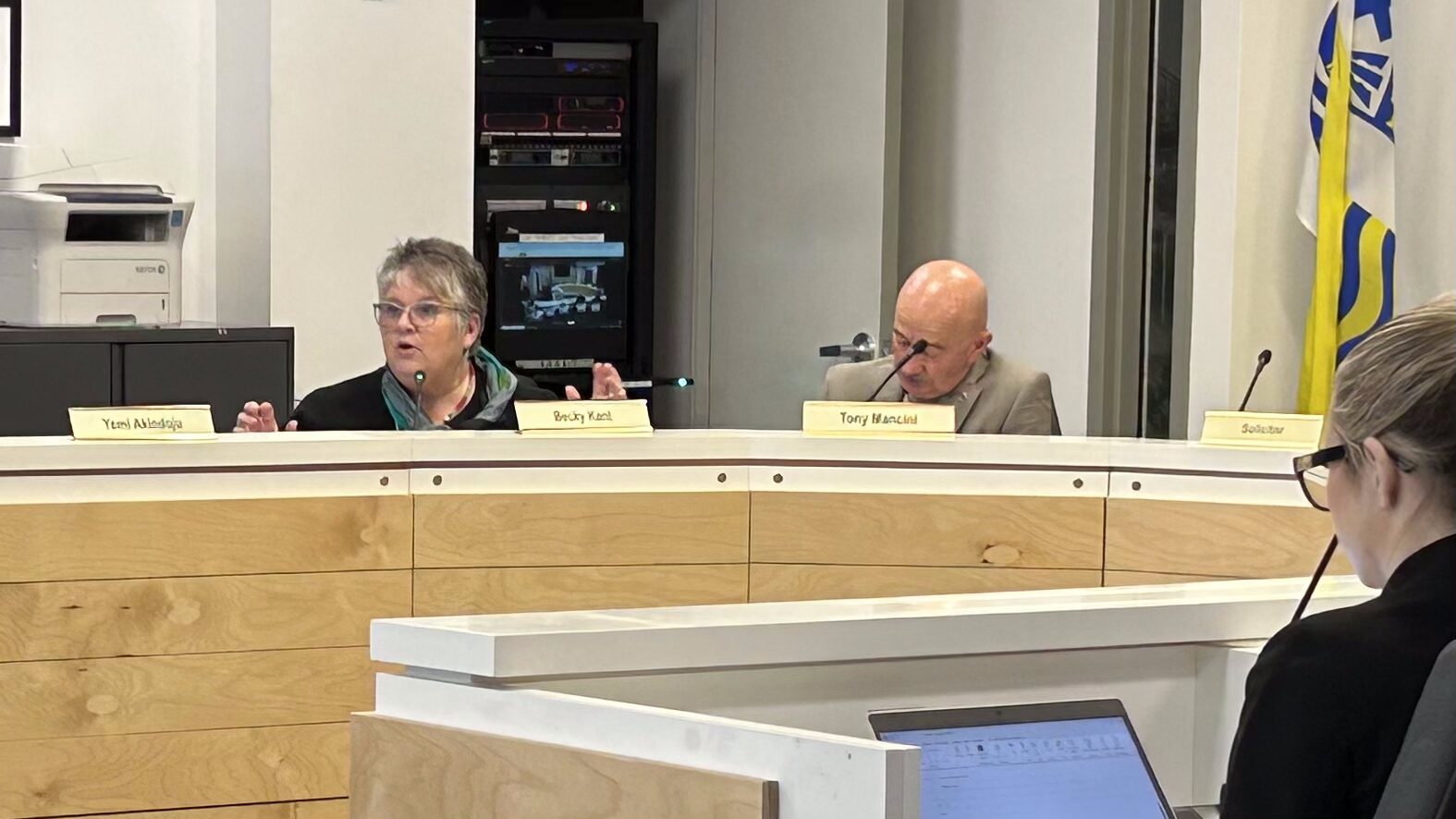 A woman sitting behind a desk talking and a man is sitting next to her with his head down.