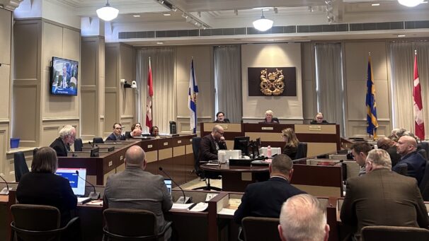 Three men sitting back on, looking at the halifax regional council sitting at a round desk.