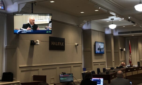 A TV projects a man talking in city hall.