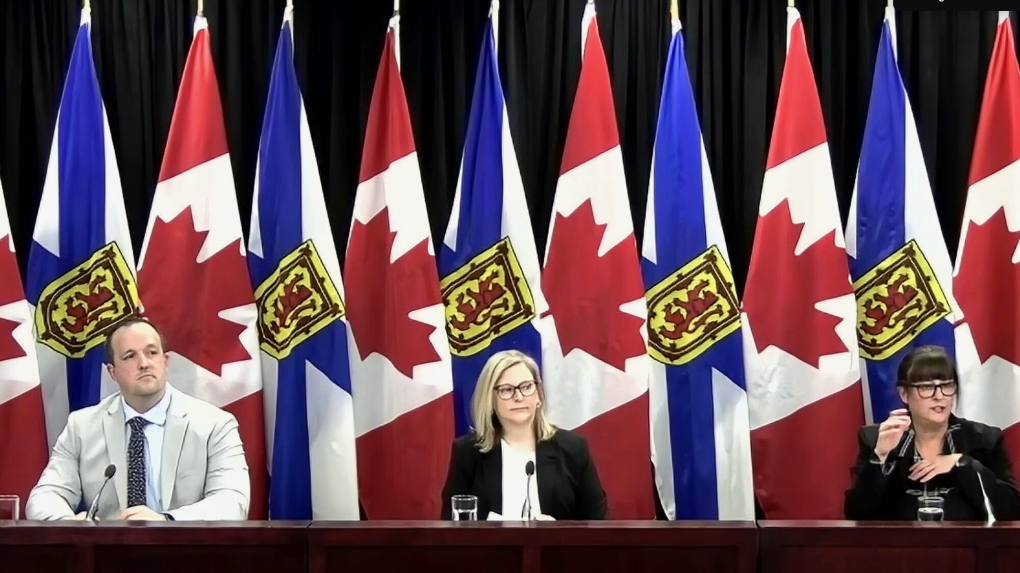 Three people sit in-front of Canadian and Nova Scotian flags for a press conference.