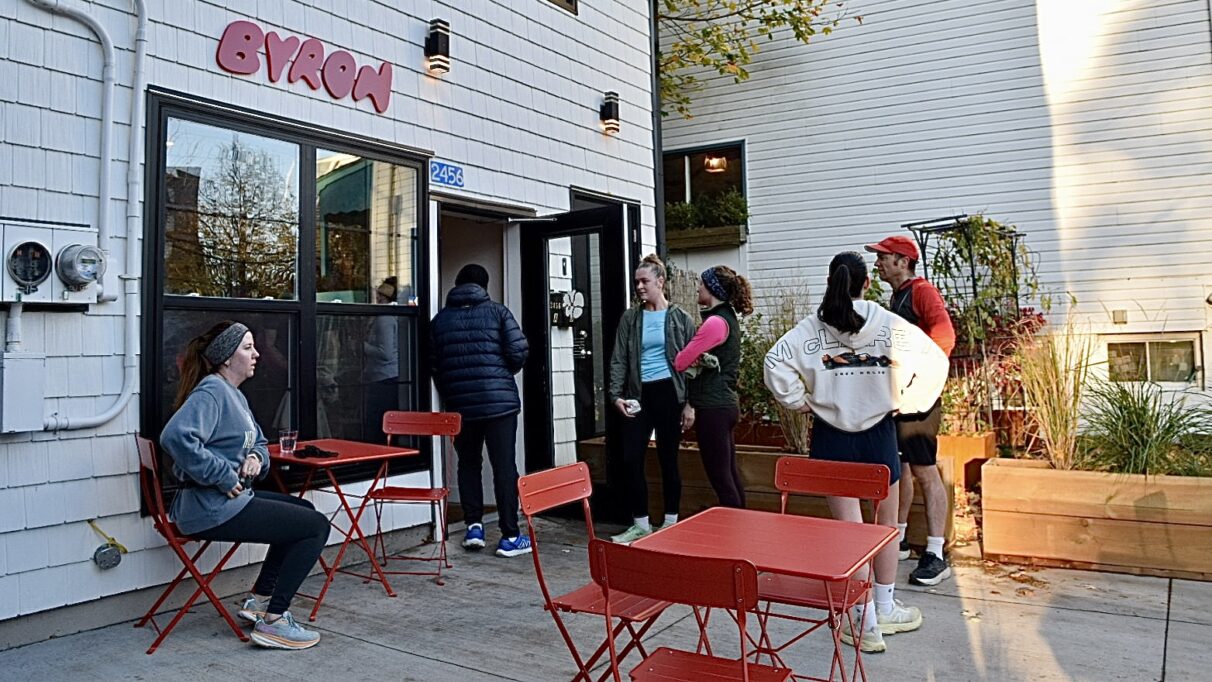 People waiting in line outside of a coffee shop; they are talking to each other.