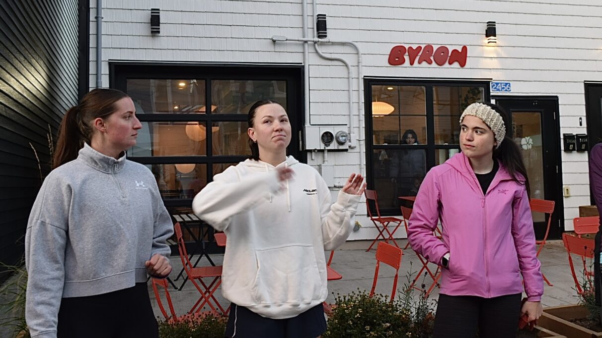 Three women standing next to each other. The women in the middle is mid conversation. The women on either side are looking at her.