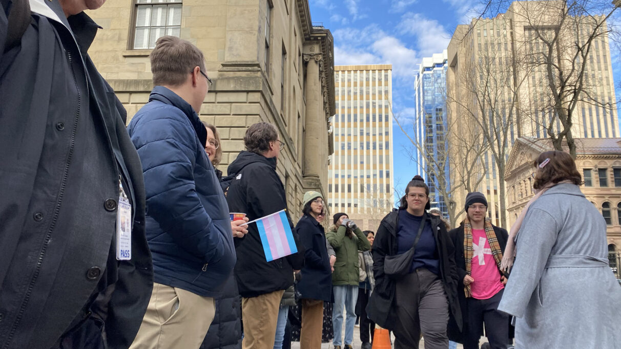 A group of people gather outside province house, one holding a trans flag