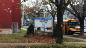 A picture of a red building to the left, with paint peeling. A sign is in the forefront and says "St. Joseph's Alexander McKay School" with lettering beneath that reads "Friendships are the best ships". The letters are askew.