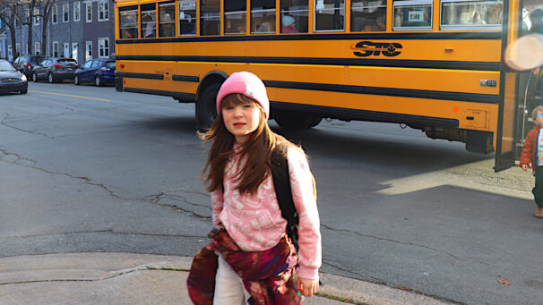 A girl in a pink sweater and pink hat walks on the sidewalk after getting off the yellow bus behind her.