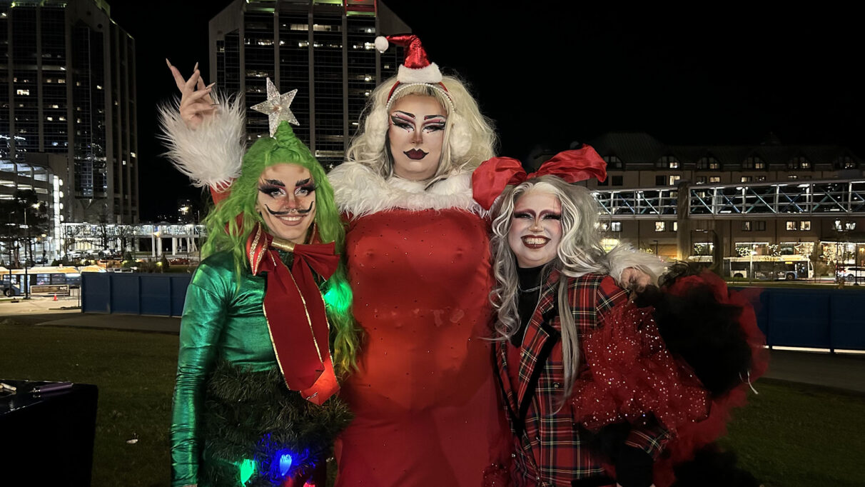 3 drag artists posing for a photo in front of buildings.
