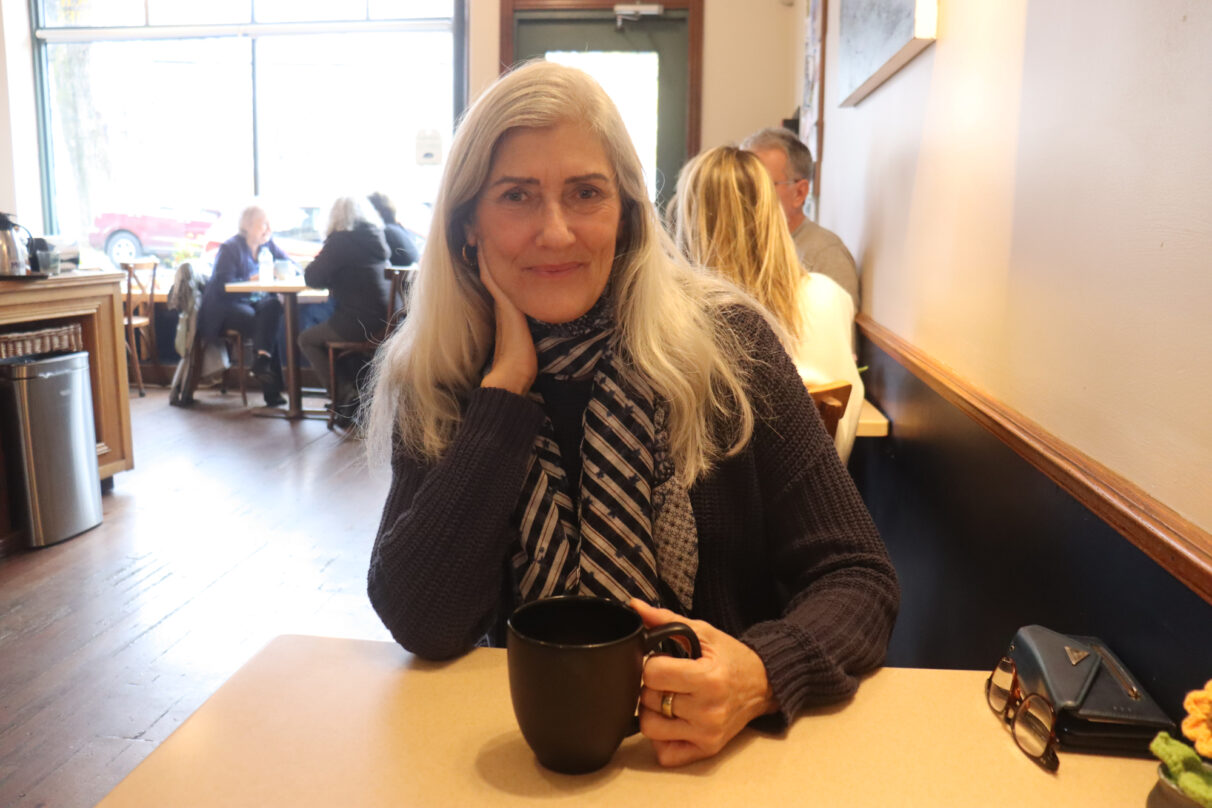 A woman with white hair smiles at the camera. One hand holds a black mug.