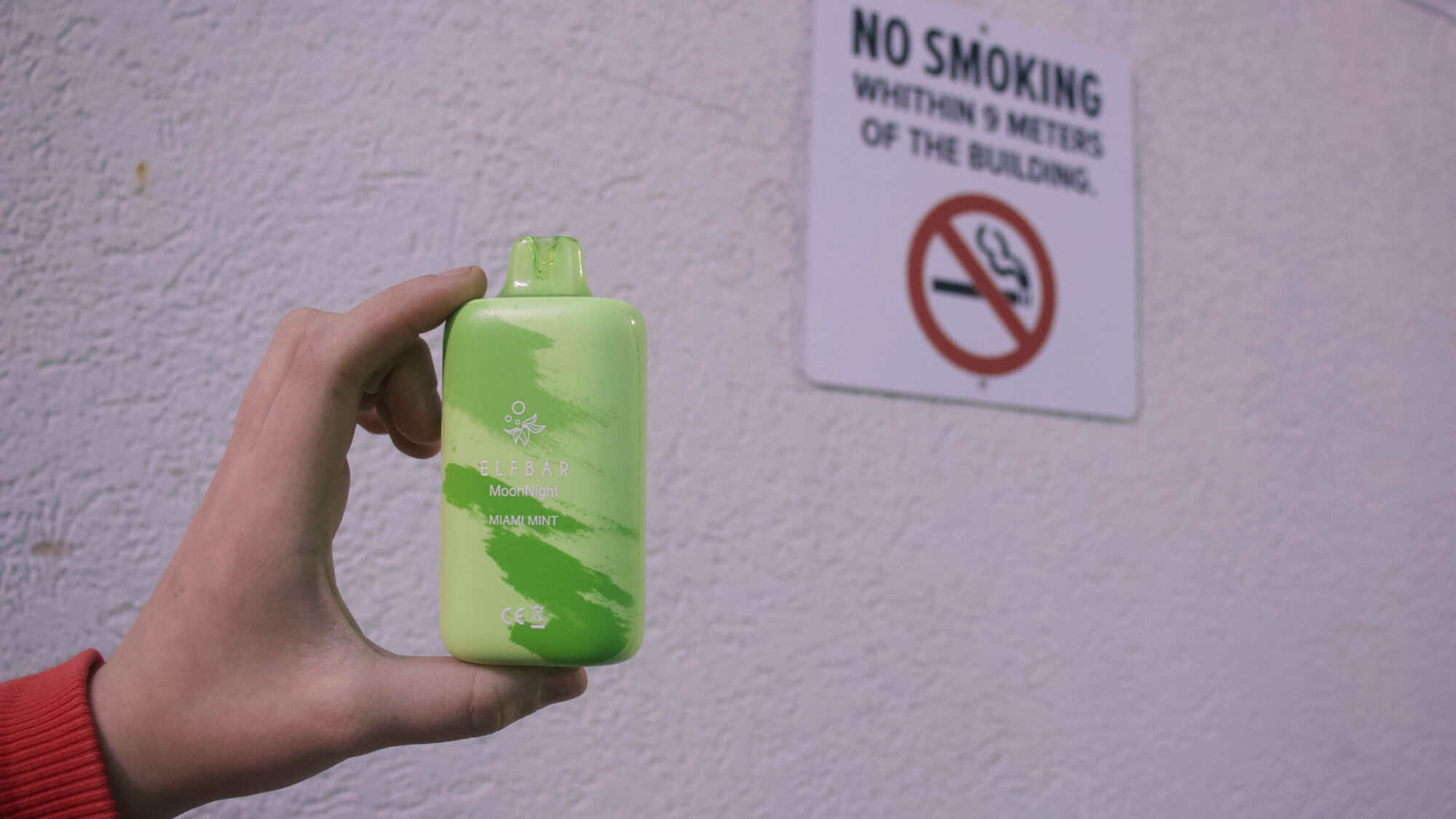 Someone holds a green “miami mint” flavoured disposable Elfbar vape in front of a “no smoking” sign.