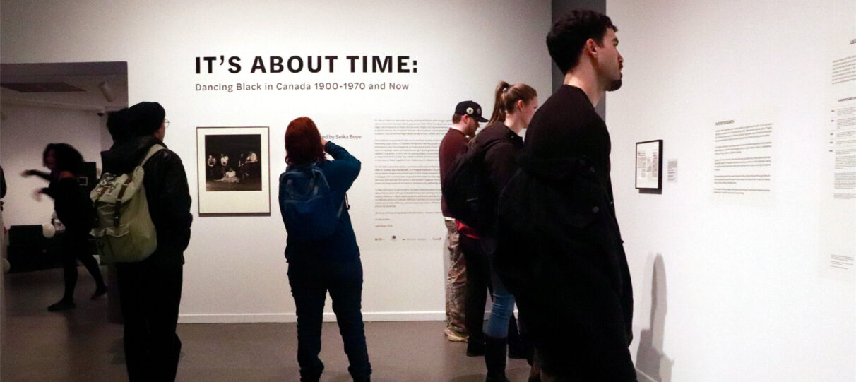 People read and take photos of art displayed on white gallery walls with the exhibition title in the background.