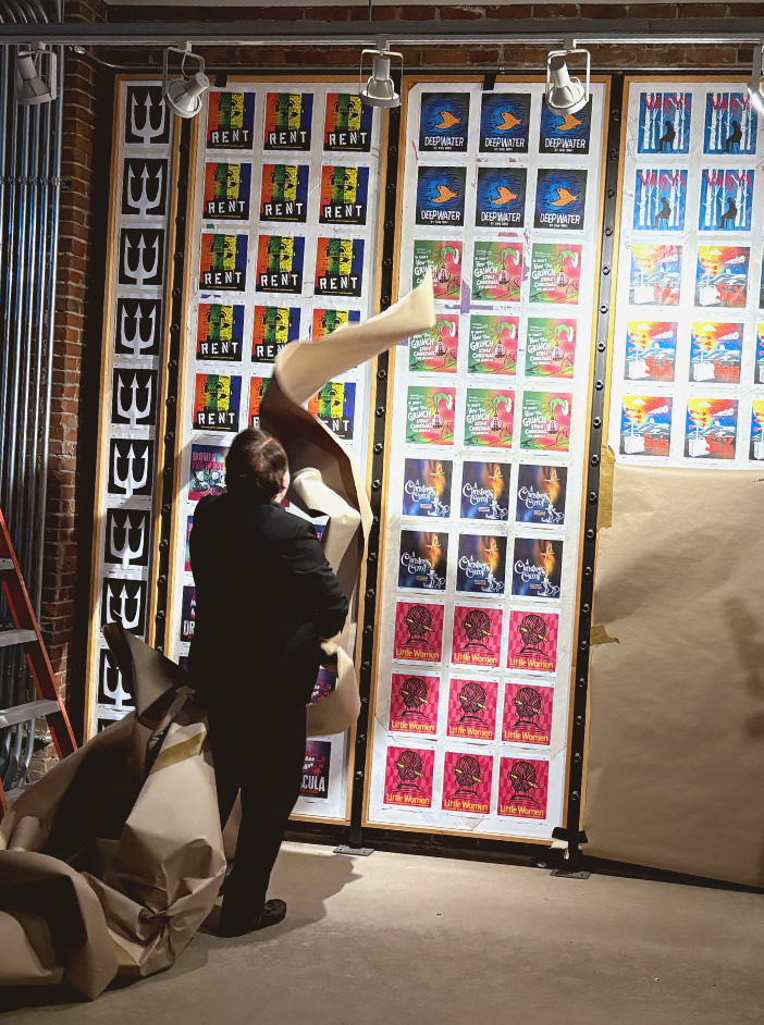 Jeremy Webb tears brown paper off a wall to reveal the posters for the Neptune Theatre 2026-2027 shows.