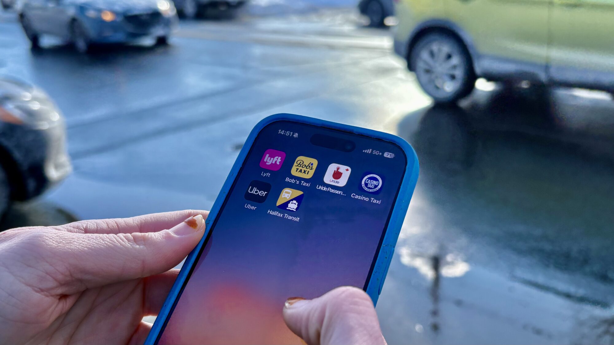 A phone screen belonging to Barley Burns shows six applications that are used to get vehicle for hire services/transportation, including Lyft, Bob’s Taxi, URide, Casino Taxi, Uber and Halifax Public Transit. The background is a street with three cars visible. The car in the top right is green. There is sunshine visible.