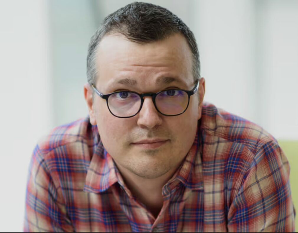 A man with glasses and a plaid shirt sitting and looking at the camera