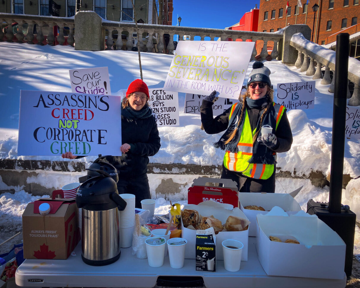 Two former Ubisoft employees hold up protest signs at the rally for Ubisoft Halifax at Grand Parade. There is a table of refreshments in front of them. The signs they are holding read “Is the ‘generous severance’ in the room with us?” and “Assassins Creed, not corporate greed”.
