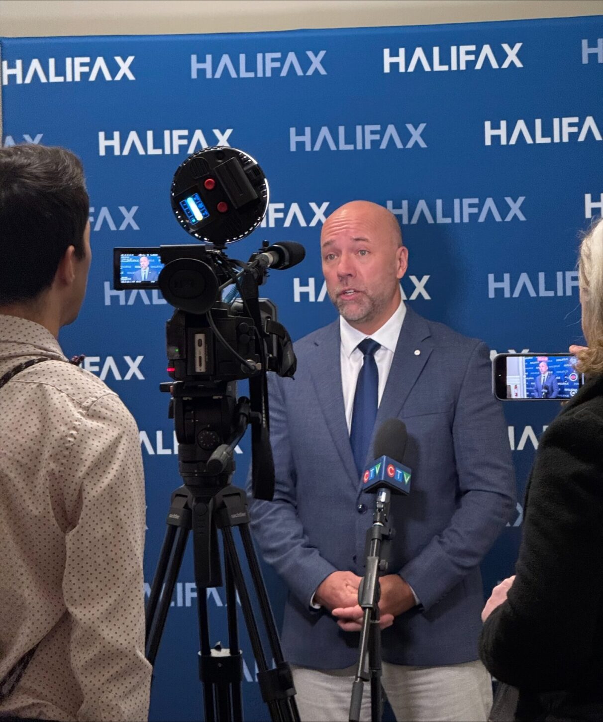 Two journalists stand with their backs to the camera, filming and recording with a video camera, mic and smartphone a statement from Halifax mayor Andy Fillmore, a middle aged bald man with a stubble beard in a blue suit jacket and blue tie. He is standing in front of a Halifax city branded backdrop.