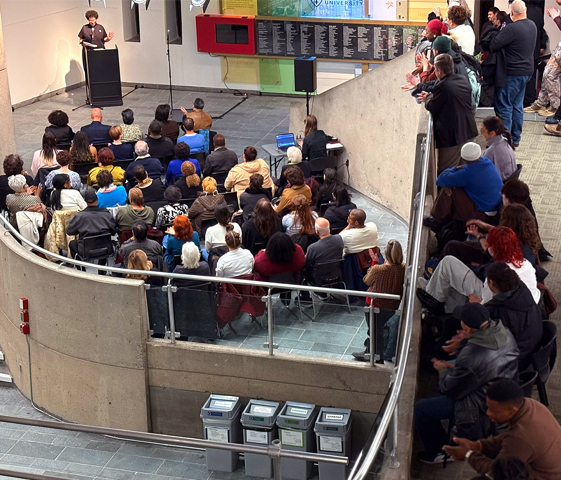 A woman at a podium speaks to guests seated and standing, with guests overlooking from the second floor.