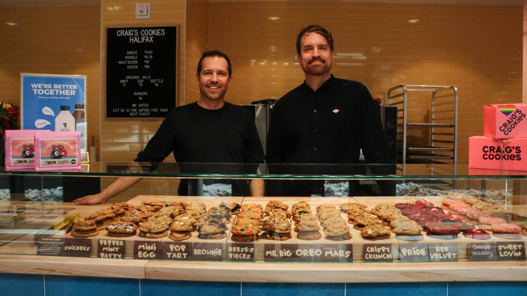Craig Pike and Brendan Waddell stand behind the counter at their new Halifax location of Craig’s Cookies. There are a number of colourful cookies and their flavours displayed in front of them.