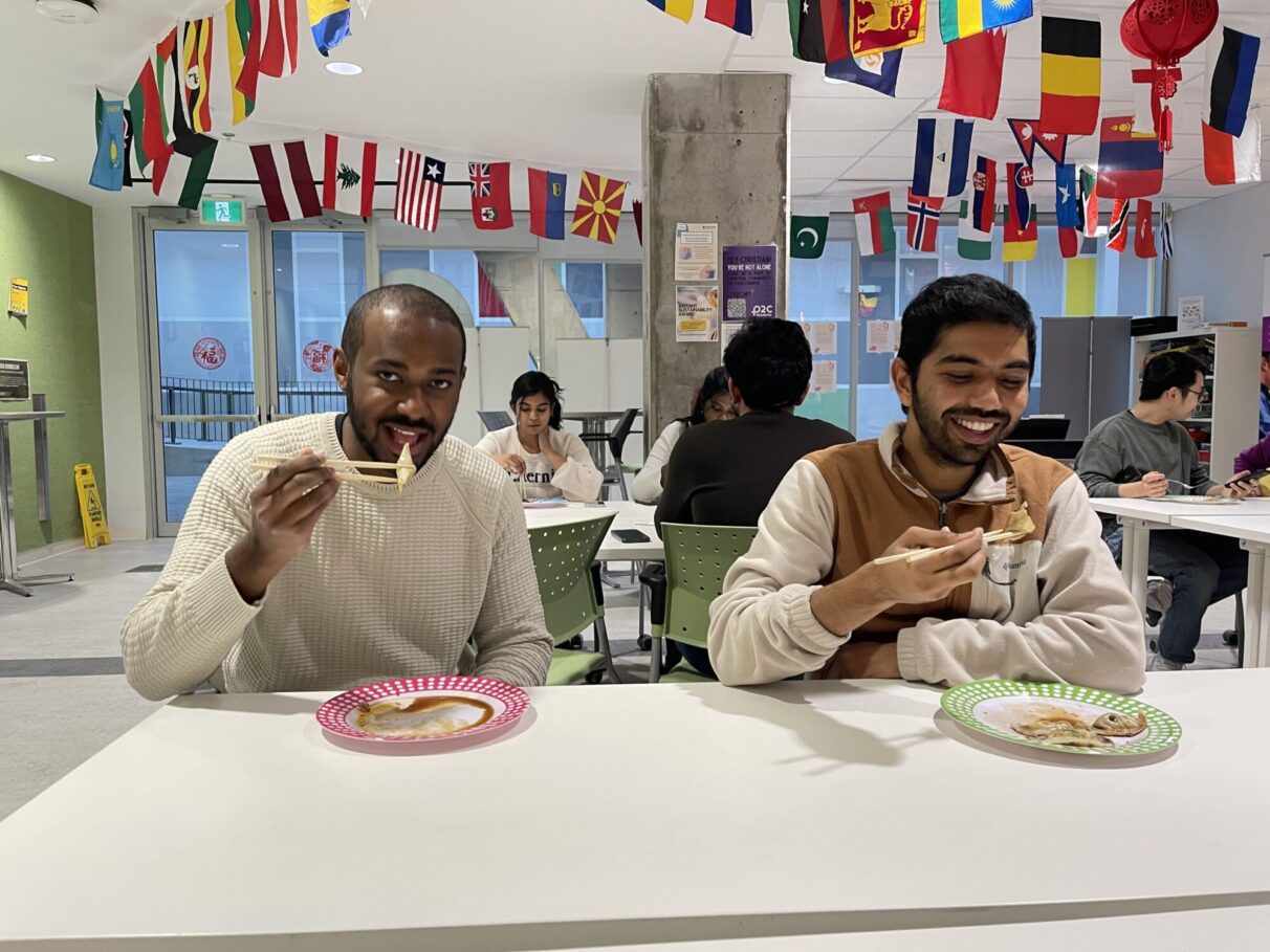 Two students looking at the camera and eating dumplings with chopsticks