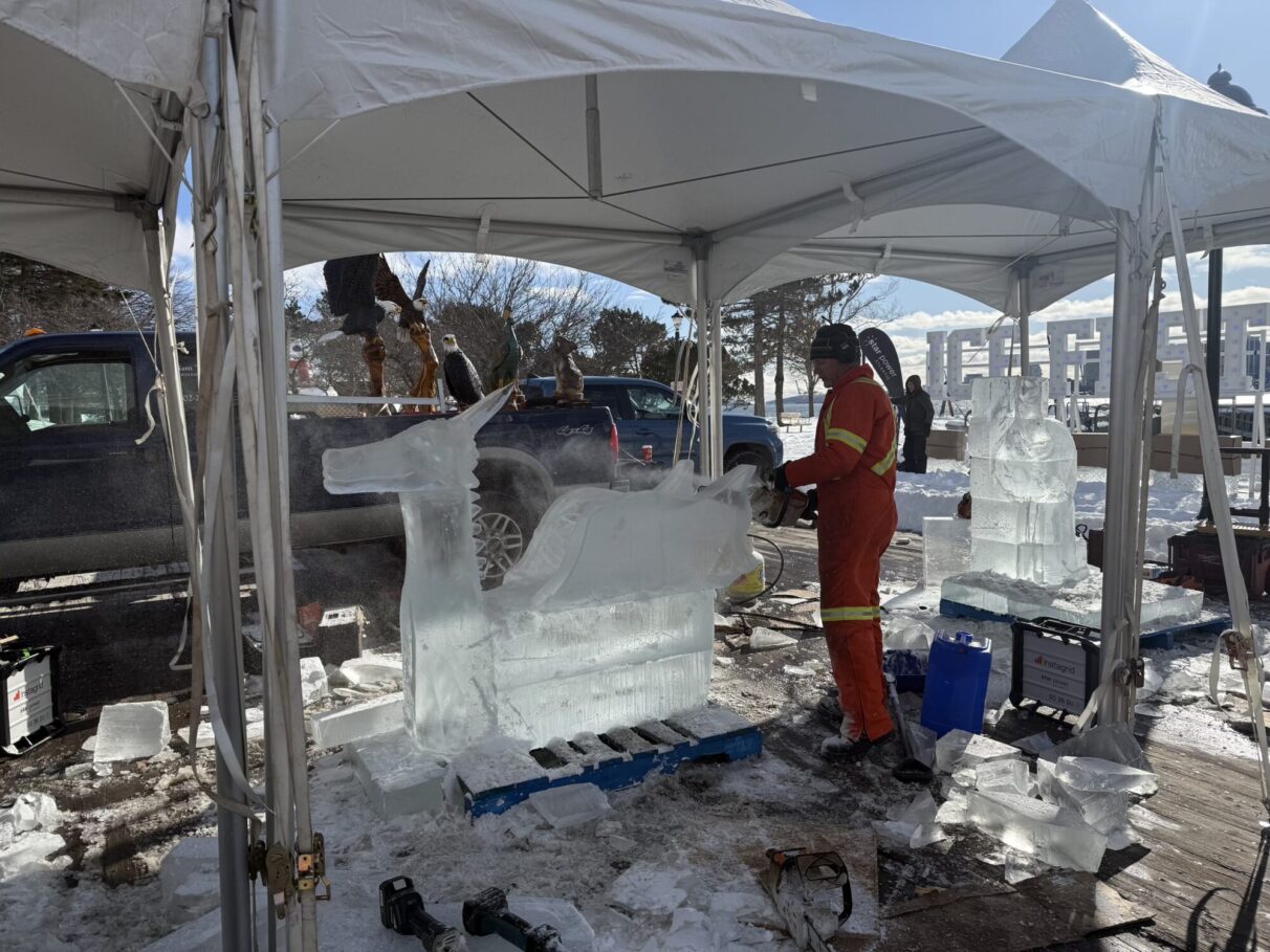 A man in orange uses a chainsaw to carve a dragon out of ice.