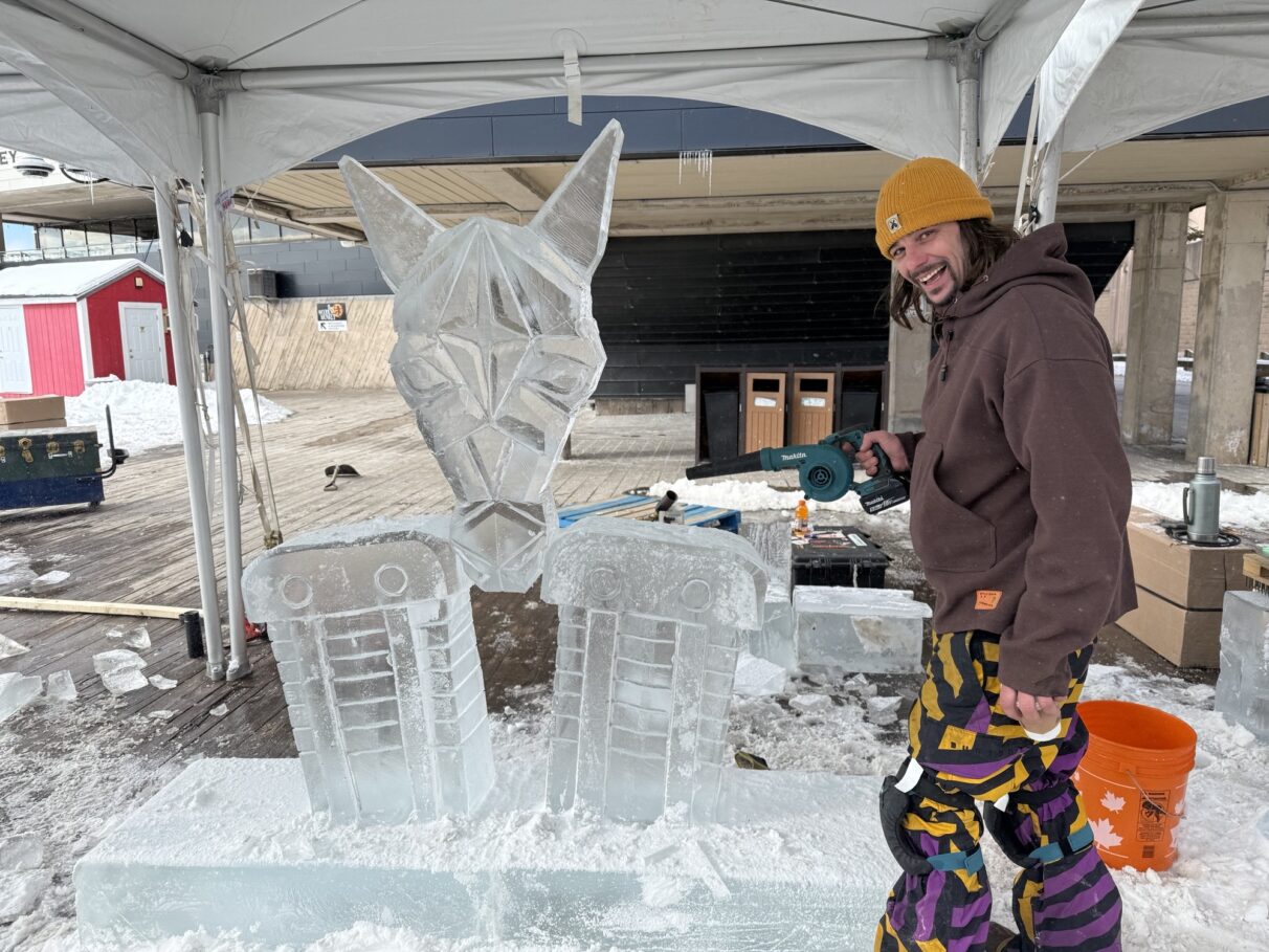 An ice sculpture featuring a horse head and two pedestals is displayed outside of the Alderney Ferry Terminal. Ice carver Joel Palmer is pictured in colourful snowpants, a brown hoodie and a yellow beanie, holding a heat blower.