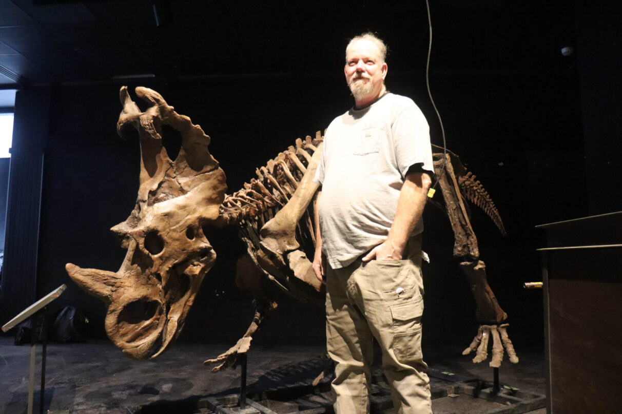 A man stands in front of a horned dinosaur skeleton.