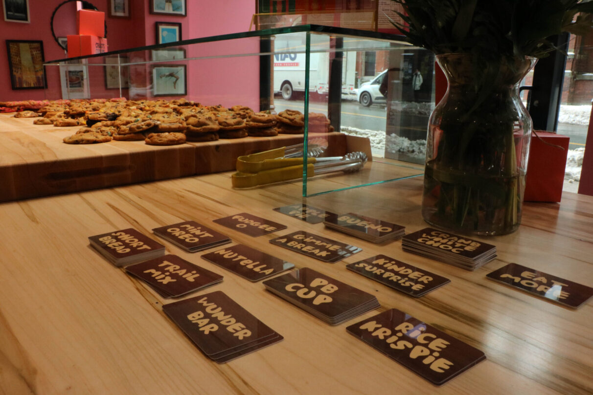 Behind the counter of Halifax’s new Craig’s Cookies location. Flavours are written on cards to be put out with cookies, and cookies appear laid out on wood beside them.