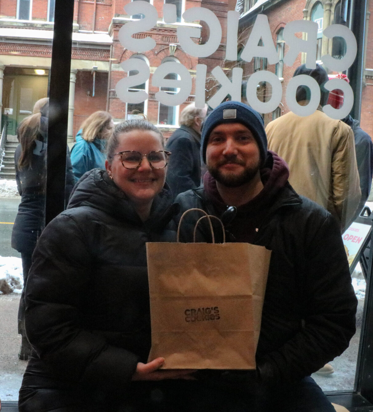 Two people sit holding a brown bag labeled Craig’s Cookies in Craig’s Cookies’ Halifax location.