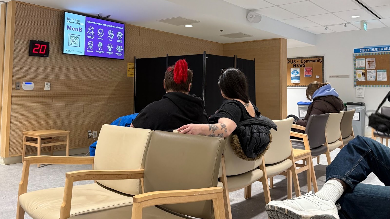 Three figures sit in chairs in a waiting room at the Dalhousie Student Health & Wellness Centre. One of the figures has a red pony tail.