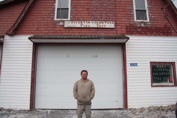 A man stands in front of a fire hall that has chipped paint and reads 'Tiverton volunteer fire dept'. The man is wearing a sweater and cargo pants.