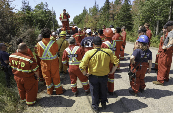 A man stands on a pick-up truck in front of a group of volunteer firefighters wearing red coveralls. They are doing a briefing on a dirt road with forest in the background.