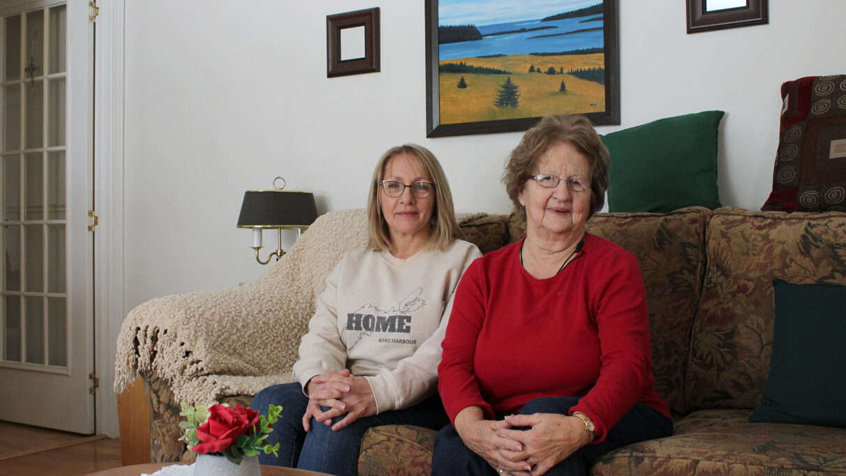 Maureen Fraser and Mary Burns sitting on their living room couch in front of a painting of Wine Harbour, NS.